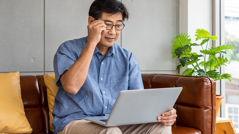 Asian man wearing and holding is glasses as he is using is laptop