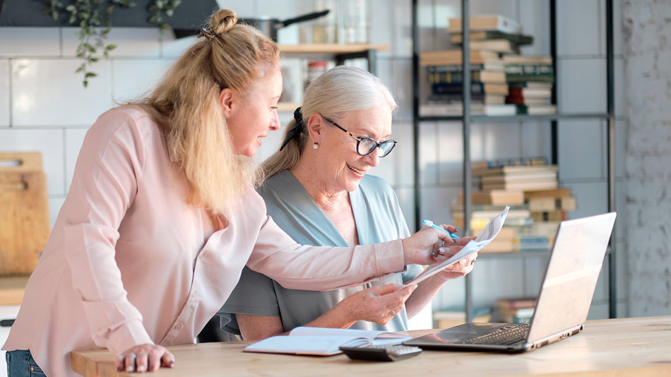 Two senior women having a discussion in the kitchen while looking at papers with a laptop open.